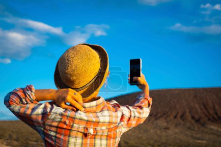Foto de Back view of woman tourist taking selfie picture in outdoor leisure adventure activity alone. One woman using mobile phone like a camera to take pictures of the landscape. Travel destination people - Imagen libre de derechos