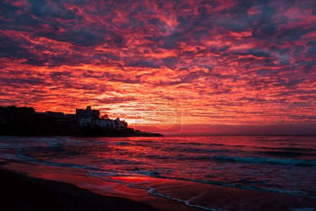 Téléchargez les photos : Red sunset at the beach with town in silhouette and dramatic sky with clouds. Amazing sunlight sunrise seascape with waves and houses in background. Concept of travel destination - en image libre de droit