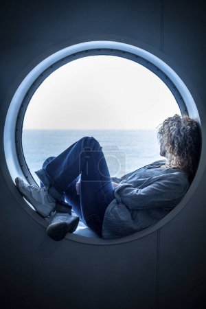 Traveler passenger woman sitting inside a porthole in the boat cruise enjoying the trip and journey travel adventure alone contemplating ocean water. Concept of transport by sea and female people