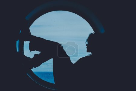 Téléchargez les photos : Silhouette d'une passagère de bateau se relaxant assis sur un grand hublot de fenêtre avec les vagues de l'océan de mer en arrière-plan à l'extérieur. Les gens en voyage mode de vie. Transport sur l'océan. La soif d'errance - en image libre de droit