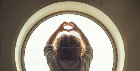 Foto de Back view of a woman doing heart sign gesture with hands in a circle window background and light outside. Concept of happiness and healthy freedom lifestyle, female people alone. Copy space - Imagen libre de derechos