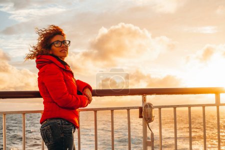 Téléchargez les photos : Tourist woman enjoy warm orange sunset on the dock of a ferry boat ship during holiday vacation travel. Happy traveler on the ocean. Sea waves and horizon in background. Journey adventure - en image libre de droit
