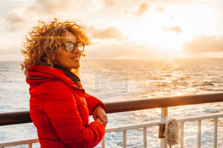 Téléchargez les photos : Portrait of a cheerful  woman enjoying sunset outside, traveling the ocean on a ferry cruise ship boat alone. Traveler people lifestyle, Red jacket. Orange warm sky background. Transport - en image libre de droit