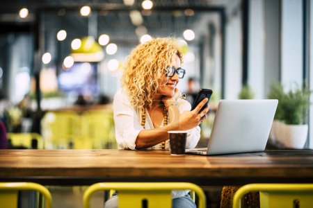 Photo for Happy adult woman using phone and laptop at the desk in cafe with free wireless connection. Modern businesswoman working online in coworking space. Smart people with online job indoor activity - Royalty Free Image