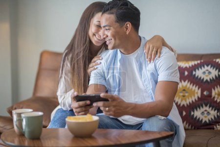 Photo for Young couple watching online content in a smartphone sitting on a sofa at home in the living room. Happy boy and girl enjoy cellphone connection together relaxing on couch. - Royalty Free Image