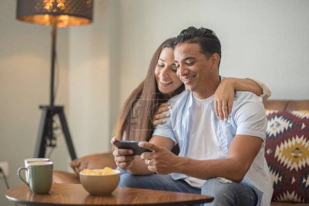 Photo for Happy young happy couple using smartphone social media apps at home, smiling husband and wife millennial users customers talking bonding watching funny video looking at mobile phone relaxing on sofa - Royalty Free Image