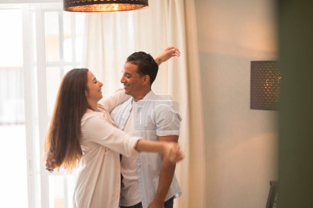 Photo for Smiling man hugging charming Caucasian woman, two people standing and joyfully looking at each other. Young international couple happily spending time in cozy modern home. Indoor leisure life - Royalty Free Image