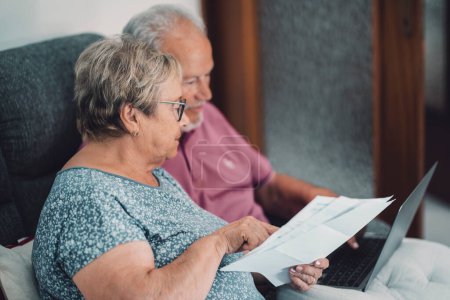 Side view of real senior couple at home paying bills online with internet banking app and account. Concept of life costs and gas energy bills for old senior people, Man and woman elderly lifestyle.
