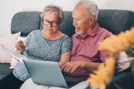 Photo for Old senior couple at home paying bills online with web banking app and checking the amounts on documents. Elderly and modern technology, real life. Mature man and woman using computer and internet - Royalty Free Image