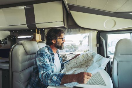 Foto de Freedom and vehicle van  lifestyle. One happy mid-age man smiling and planning road's trip on a map guide sitting inside a camper van motor home. Alternative cozy nomadic home and life. Vacation. - Imagen libre de derechos