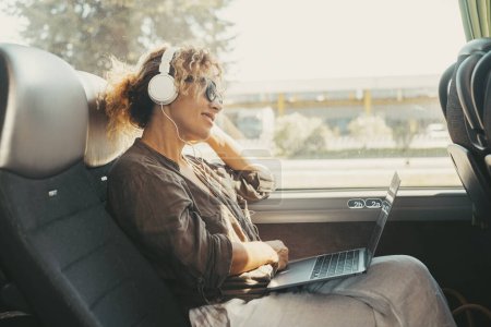 Foto de Modern travel lifestyle people with happy smiling woman listening music with headphones and laptop computer sitting and relaxing on a bus seat as a travel passenger. Transportation female people - Imagen libre de derechos