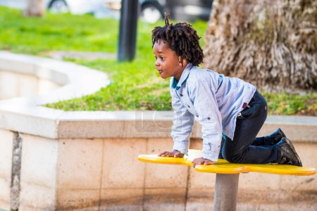 Téléchargez les photos : Jeune enfant afro-américain profiter du temps de s'amuser à l'aire de jeux dans le parc de la ville. Petit gars jouant dans les loisirs de plein air seul - en image libre de droit