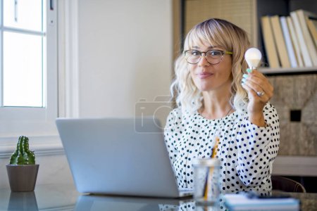 Photo for Businesswoman holding light bulb on the desk in office and using computer. Corporate portrait of home worker female people having idea. Concept of creativity and solutions. Successful businesswoman - Royalty Free Image