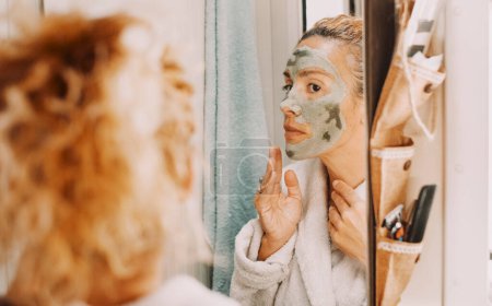 Téléchargez les photos : Portrait miroir de femme adulte d'âge moyen utilisant la crème verte sur son visage comme routine quotidienne de bien-être pour les rides et les soins de la peau. Style de vie naturel et traitements sur les femmes. Vous aimer dame - en image libre de droit