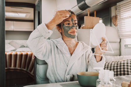 Photo for Portrait of real adult woman applying green mud beauty mask on face using mirror and hands inside a camper van. Alternative lifestyle freedom people travel and nature. Daily routine - Royalty Free Image