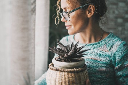 Photo for Side view of one pensive woman holding a plant inside the home and looking outside the window alone. Female people wearing eyewear and caring interior garden nature. Serene people feeling emotion. Sadness - Royalty Free Image