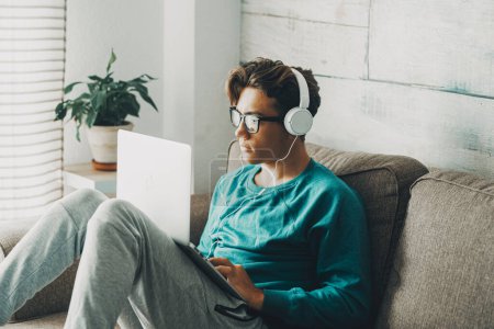 Photo for Relaxed young teenager using laptop sitting on the sofa at home for play or study. Wearing headphones. One teenager boy listening music in indoor relax leisure activity alone. Afternoon time alone - Royalty Free Image