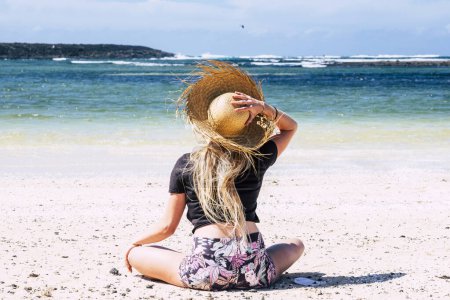 Foto de Vista trasera de la joven mujer disfrutando del sol en la playa en la isla tropical y el destino de vacaciones de verano. Baño de sol. Chica turista con sombrero de paja ocio al aire libre actividad de arena wanderlust - Imagen libre de derechos