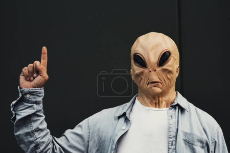 Téléchargez les photos : Portrait de masque extraterrestre homme sur un fond de mur vert pour écrire votre texte. Concept d'image pour les ventes en magasin offre de réduction avec la personne pointant la main du doigt et regardant la caméra - en image libre de droit