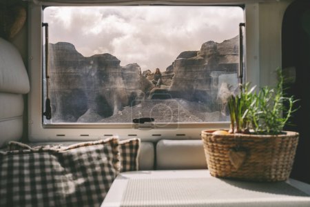 Foto de Mujer sentada en las rocas vista desde el interior moderna caravana a través de la ventana. Concepto de gente de estilo de vida nómada y destino turístico escénico. - Imagen libre de derechos