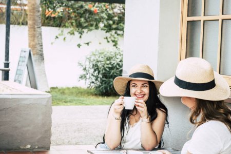 Téléchargez les photos : Jeune femme prenant un café avec un ami et sourire. Heureux touristes dans les loisirs de plein air boire cappuccino dans l'amitié. Les filles ensemble avoir du plaisir à porter des chapeaux de paille. - en image libre de droit