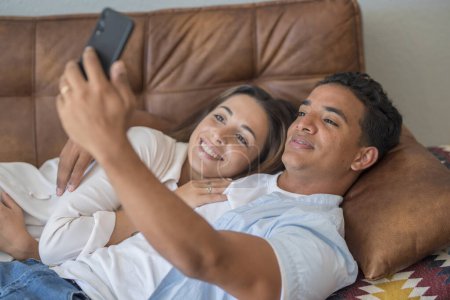 Photo for Dating and relationship. Young couple interracial black boy and caucasian girl enjoying time at home in indoor leisure activity using mobile phone to take selfie together picture laying on the sofa - Royalty Free Image