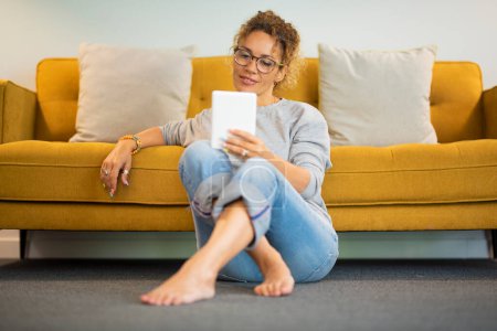 Photo for Easy relaxed lady at home reading and using tablet, ebook device alone witting on the ground against the sofa. Relaxation and independent lifestyle female people. Young adult lady in indoor - Royalty Free Image