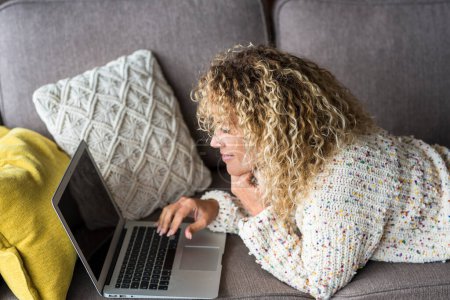 Photo for Woman using laptop comfortably laying on the sofa at home smiling and enjoying wireless technology. House working leisure activity. Female people surfing the net and having relax indoor - Royalty Free Image