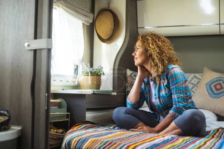 Photo for Travel lifestyle and dreaming people destination. One serene happy woman sitting on bed inside camper van bedroom and looking outside the window with dreamer expression. Freedom and independence life - Royalty Free Image