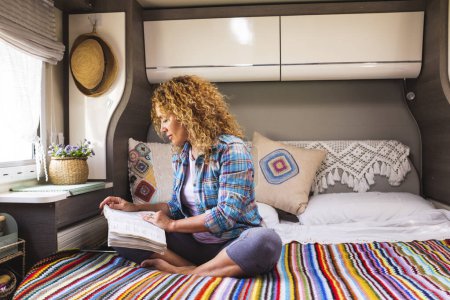 Photo for One woman reading a book comfortably sitting on bed inside a camper van modern motor home alternative, tiny cozy house. Relaxation and indoor leisure activity people lifestyle. Travel and van life lady - Royalty Free Image