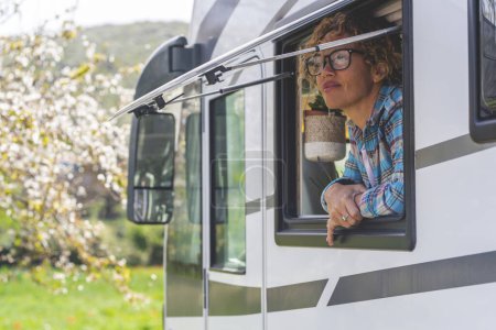 Photo for Travel vacation camper van lifestyle. One happy and free alone woman having relax looking outside the window of her modern camper van motor home. Journey adventure holiday trip. Nature and free parking - Royalty Free Image