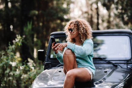 Téléchargez les photos : Femme adulte assis sur le devant de la voiture et en utilisant un téléphone portable avec forêt bois vert autour. Les gens qui aiment voyager et le véhicule dans la nature à l'extérieur. Voyageur de véhicule, femelle utilisant la connexion - en image libre de droit