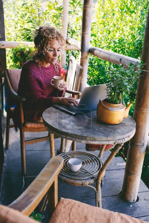 Téléchargez les photos : Femme sereine saine assise à la maison en plein air dans un patio avec fond vert de la nature et à l'aide d'un ordinateur portable avec connexion sans fil. Concept de bureau et d'emploi alternatifs. - en image libre de droit