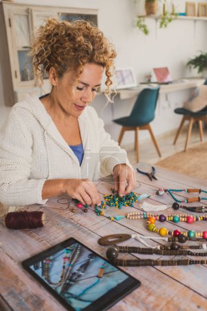 Photo for Happy woman working with beads and cords to make necklace and accessories, learning to watch online tutorial on tablet with internet connection. Indoor hobby leisure activity. New job concept lady - Royalty Free Image