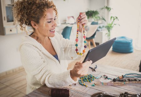 Photo for Happy woman indoor leisure hobby work activity producing beads accessories necklaces showing her jewel in video call using tablet and wireless connection technology. New business at home lifestyle - Royalty Free Image
