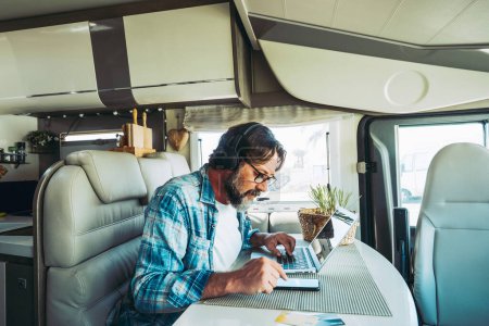 Photo for Man using computer and phone sitting indoor inside camper van modern vehicle. People enjoying technology and connection in travel vacation lifestyle. Living vanlife and working. New job office - Royalty Free Image