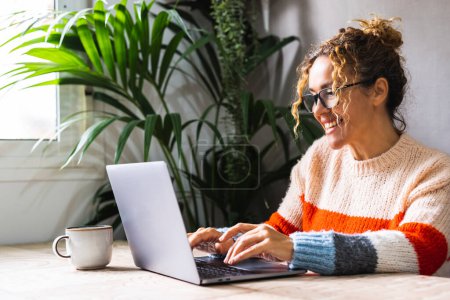 Photo for Happy woman writing on laptop on desk table in home office workplace. Adult female cheerful portrait wearing glasses and using laptop smiling. Modern alternative business job entrepreneur business - Royalty Free Image