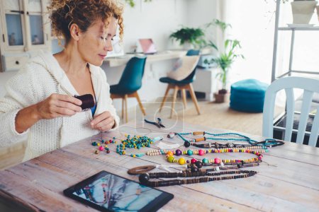 People in indoor leisure activity concept lifestyle.  woman doing beads necklace on the table in living room with tablet device on. Modern alternative new business work online selling on web