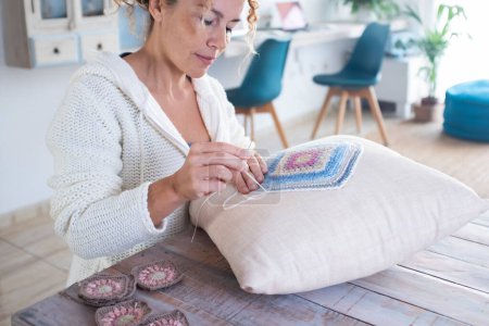 Portrait of happy serene woman working embroidery on a white pillow in living room at home sitting at the table and smiling. Renewal house decoration. Indoor leisure activity hobby at home. Art craft
