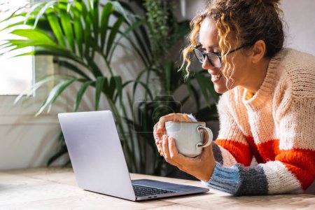 Photo for Modern woman smiling at the laptop display and enjoying time online alone. Video call communication concept. Happy female people using computer notebook at home on the table and holding tea cup - Royalty Free Image