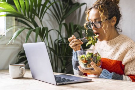 Photo for Woman on diet eating fresh salads from transparent bowl while using laptop at home watching online content. Young entrepreneur busy in new business concept lifestyle online. Lunch break time lady - Royalty Free Image