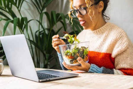 Photo for Woman eating a salad with disgusted expression. People on diet and healthy lifestyle habits. Healthy food nutrition concept. Lady tired to eat vegetables. Modern female using computer and eat lunch - Royalty Free Image