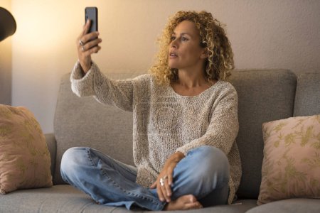 Photo for Woman using cellphone at home having relax on sofa alone. Female people taking selfie or doing videocall online with mobile phone. Indoor leisure activity people. Relaxation - Royalty Free Image
