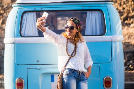 Photo for Woman taking selfie picture with phone against a blue classic van. Travel and hippy lifestyle people concept. One traveler female enjoying destination. Wearing trendy casual clothes - Royalty Free Image