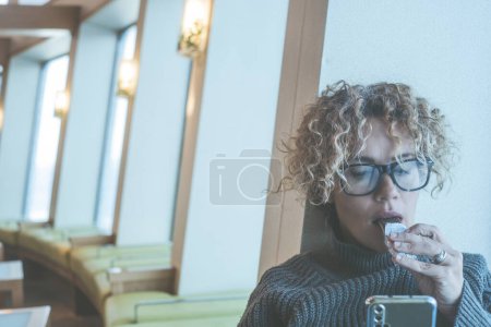 Foto de Retrato de una mujer comiendo bocadillos durante el viaje en barco y utilizando el teléfono celular para navegar por la web. Copie el espacio a la izquierda con el fondo interior. Llevando gafas. Crucero interior momento aburrido. Gente comiendo - Imagen libre de derechos