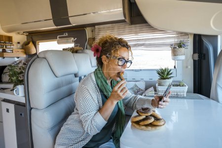 Photo for Bored woman eating a lot of donuts sitting at the table inside a camper van during travel lifestyle. Female people enjoy vacation in motorhome having relax and using mobile phone and connection - Royalty Free Image