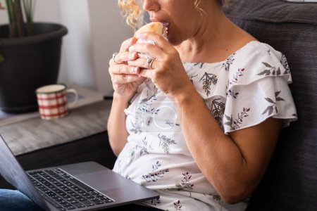Téléchargez les photos : Femme manger tout en utilisant un ordinateur portable assis sur le canapé à la maison. Femme avec nutrition sandwich. Concept de régime amaigrissant. Activité intérieure avec ordinateur et déjeuner ou collations rapides. Dame non reconnaissable - en image libre de droit