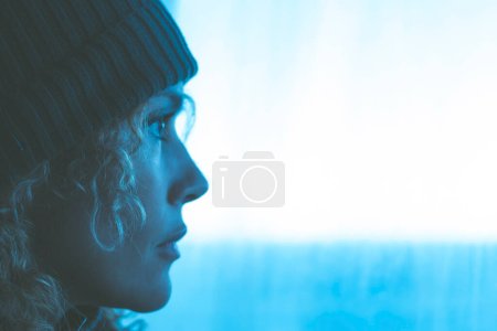 Téléchargez les photos : Portrait latéral de femme avec chapeau tricoté regardant à l'extérieur de la fenêtre en lumière bleue. Voyage aventure concept gens style de vie. Femme adulte rêvant et pensant avec fond blanc d'espace de copie. Réfléchi - en image libre de droit