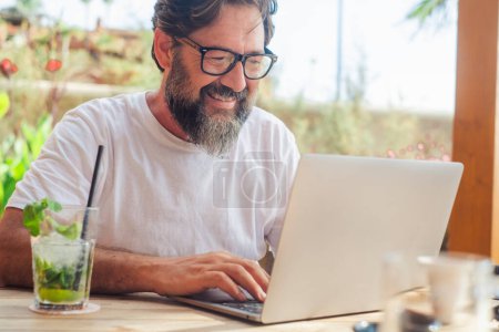 Photo for One happy mature man with beard and eyewear working with laptop in outdoor bar cafe. People enjoying roaming and wireless connection to work in alternative free office. Modern job lifestyle concept - Royalty Free Image