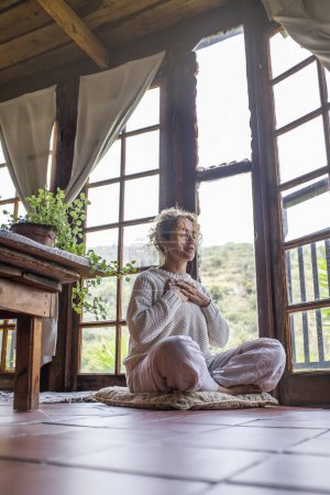 Téléchargez les photos : Femme en méditation de yoga aimez-vous position à la maison avec les mains sur la poitrine et les yeux fermés assis sur le sol. Exercice de bien-être mental sain. Soins du corps femmes. routine quotidienne intérieure - en image libre de droit